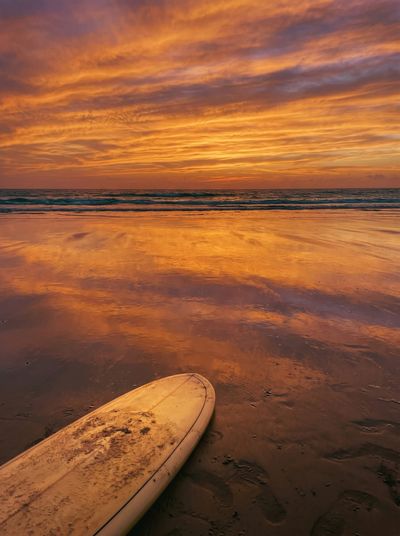 The nose of a surfboard lying on wet sand at sunset, the orange sky reflected in the shallow water