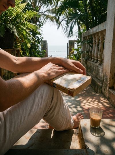 A person's hands holding a worn paperback across their knees on a balcony, a glass of chai on the ledge and the sea beyond