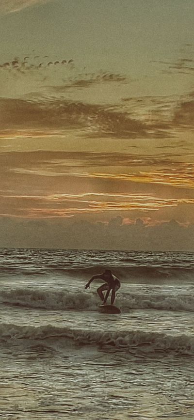 A surfer riding the white water in towards the beach at dusk under an orange and grey sky