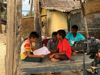 Four boys sitting on a wooden platform under a coconut tree, one reading from a notebook, filmed by a camera on a tripod in the foreground