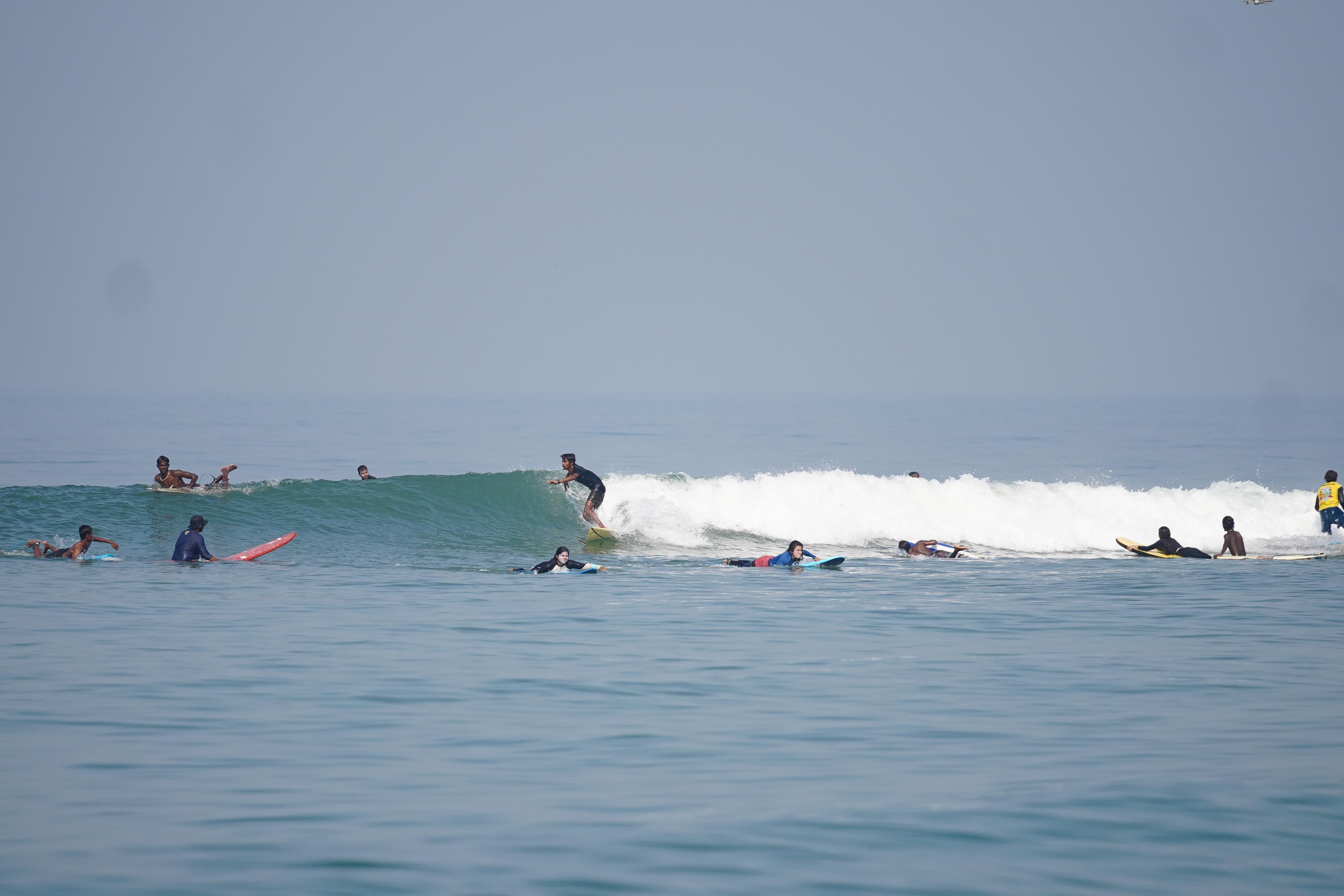 A wide line-up at Panambur, a dozen surfers sitting on their boards while one rides a small wave