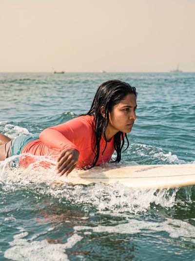 A woman lying on a surfboard in the ocean in an orange rash guard, paddling with boats on the horizon behind her