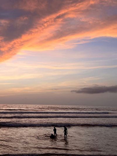 A dramatic pink and orange sunset sky filling most of the frame, three small silhouetted figures standing in the shallows below