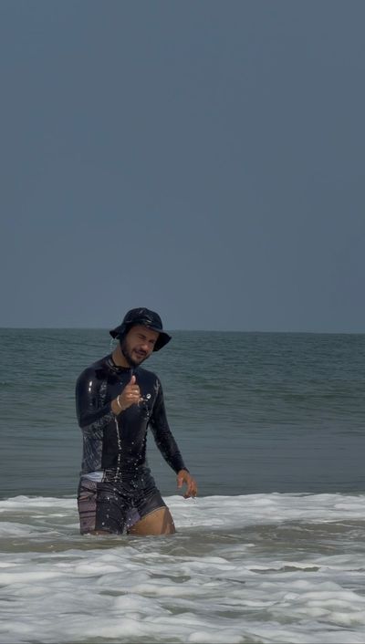 Rohan standing waist deep in the shallows in a black rash guard and bucket hat, one thumb up