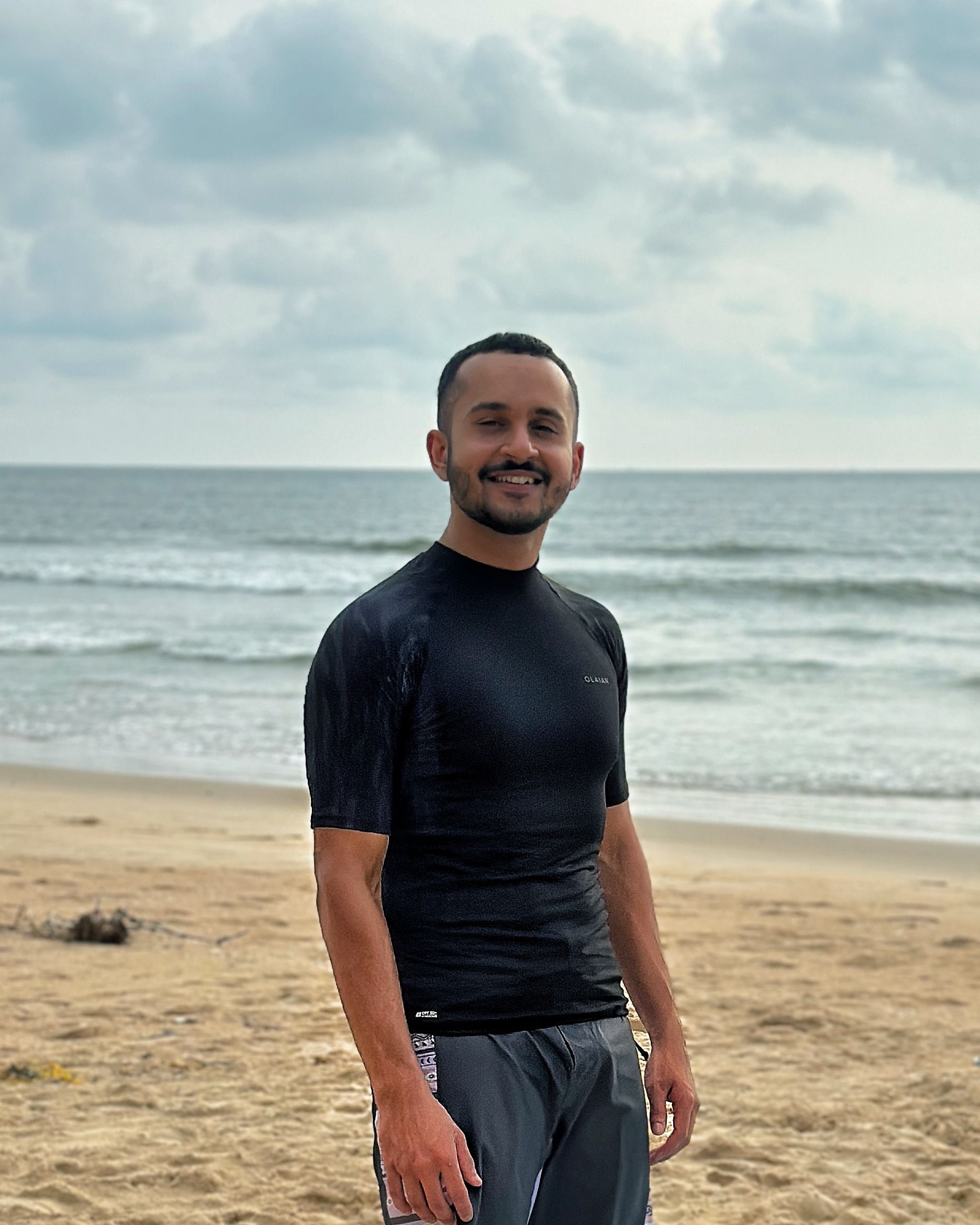 Rohan on the sand in a black rash guard, half turned and smiling, the shore break and the horizon behind him