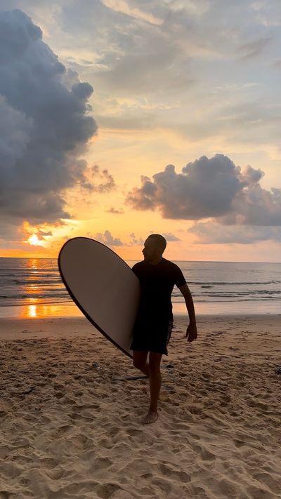 A man in a black t-shirt walking along the beach at sunset carrying a longboard under his arm, dramatic storm clouds lit orange and pink overhead