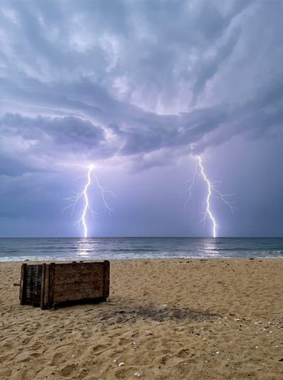 Two lightning strikes over the sea under a storm sky, a wooden crate with painted lettering sitting on the sand in the foreground