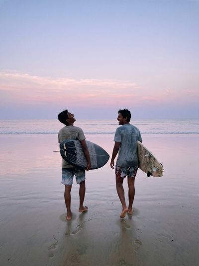Two men walking away down the beach at dusk under a pink sky, each carrying a surfboard, looking back at each other and laughing